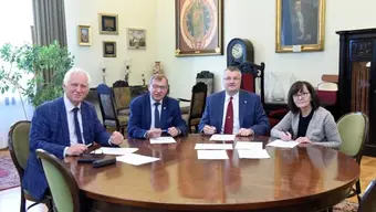 Cztery osoby siedzące przy okrągłym drewnianym stole, podpisujące dokumenty w eleganckim pomieszczeniu z obrazami, fotografiami i zabytkowymi dekoracjami