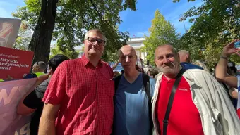Trzech związowców na tle zielonych drzew i flag.