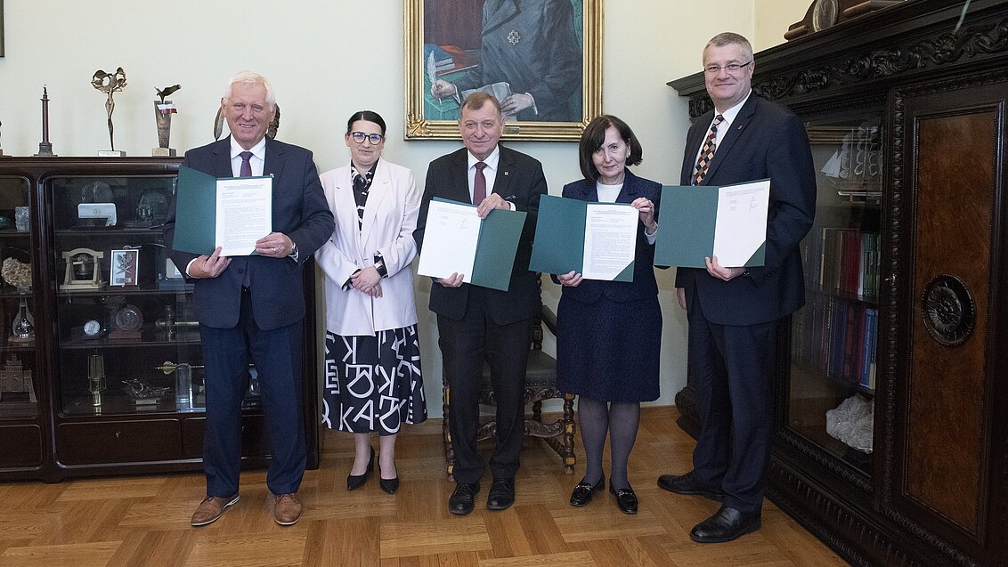 Pięć osób stoi na tle portretu Stanisława Staszica i prezentuje teczki z porozumieniem