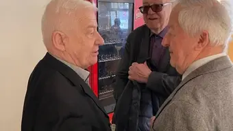 Trzech starszych mężczyzn rozmawia w pomieszczeniu; w tle widoczny automat z napojami i znak 'WYJŚCIE'. Wszyscy ubrani są elegancko, co sugeruje formalne spotkanie.