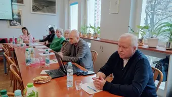 Grupa osób siedzi przy długim drewnianym stole w jasnym pomieszczeniu z dużymi oknami; na stole znajdują się napoje, szklanki, laptop i notatki. W tle widoczne są rośliny doniczkowe oraz ramki ze zdjęciami i certyfikatami na ścianie.