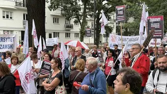 Grupa osób uczestniczy w proteście w Warszawie, trzymając transparenty z hasłami dotyczącymi sprawiedliwego wynagrodzenia i warunków pracy. Widoczne są flagi z logo organizacji oraz banery z nazwami instytucji, m.in. Uniwersytet Warszawski i Oddział Wrocław. W tle znajdują się budynki mieszkalne