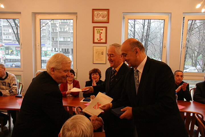 Wręczenie odznaki, dwóch mężczyzn, jeden przypina drugiemu do klapy marynarki odznaczenie