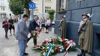 Uroczystość upamiętniająca przy budynku z tablicą pamiątkową. Dwie osoby składają wieńce z biało-czerwono-niebieskimi wstążkami, a po bokach stoją wartownicy w mundurach. W tle widoczni obserwatorzy, znak ‘TAXI’, drzewa i inne budynki.