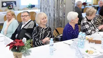 Duża sala udekorowana srebrnymi girlandami i balonami z cyfrą 2, w której odbywa się uroczystość. Goście siedzą przy długich stołach zastawionych talerzami, szklankami i napojami.