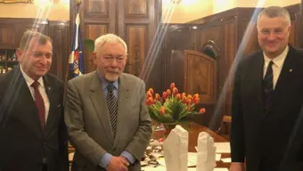 Trzy osoby w garniturach stoją w eleganckim pomieszczeniu z drewnianą boazerią; na stole przed nimi znajdują się dwie białe rzeźby i wazon z czerwonymi tulipanami, w tle widoczna flaga i ramki ze zdjęciami.