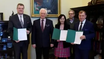 Pięć osób stojących w pomieszczeniu podczas oficjalnej uroczystości. Trzy z nich trzymają zielone dokumenty. W tle widoczny duży portret starszego mężczyzny z książką oraz drewniane meble z gablotami.